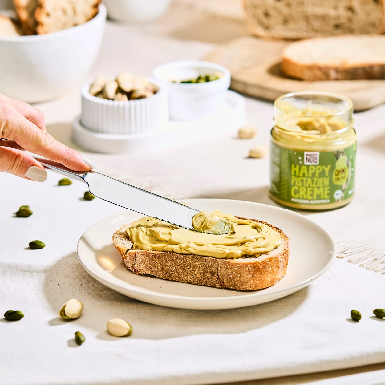 Hand streicht Happy Pistazien Creme auf eine Scheibe Brot, daneben ein Glas Pistaziencreme und Pistazien
