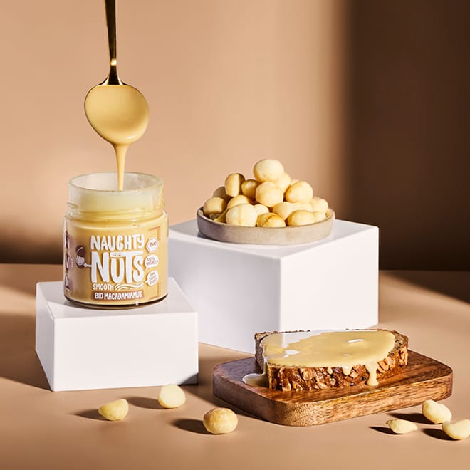 Bio Macadamiamus Smooth in Glas, Löffel, Macadamianüsse in Schale und auf Holzbrett mit Brot