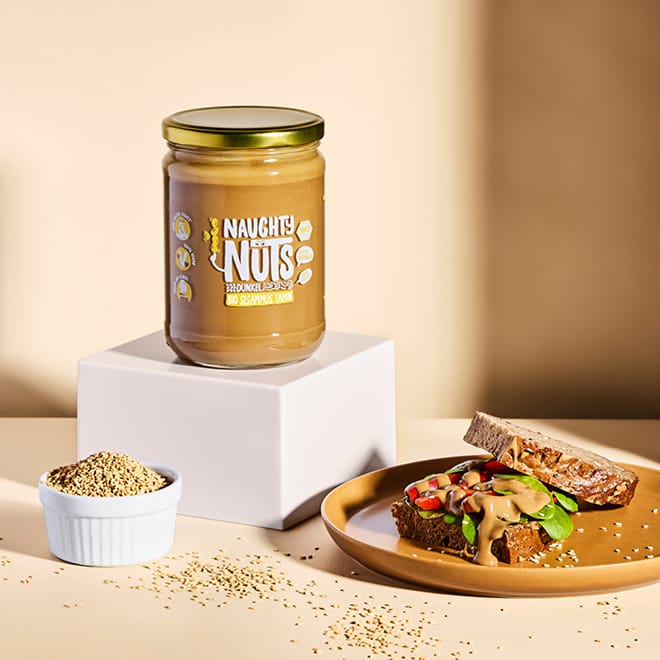Bio Sesammus Tahin dunkel (500g Glas) auf einem weißen Podest mit Sesamsamen und Brot
